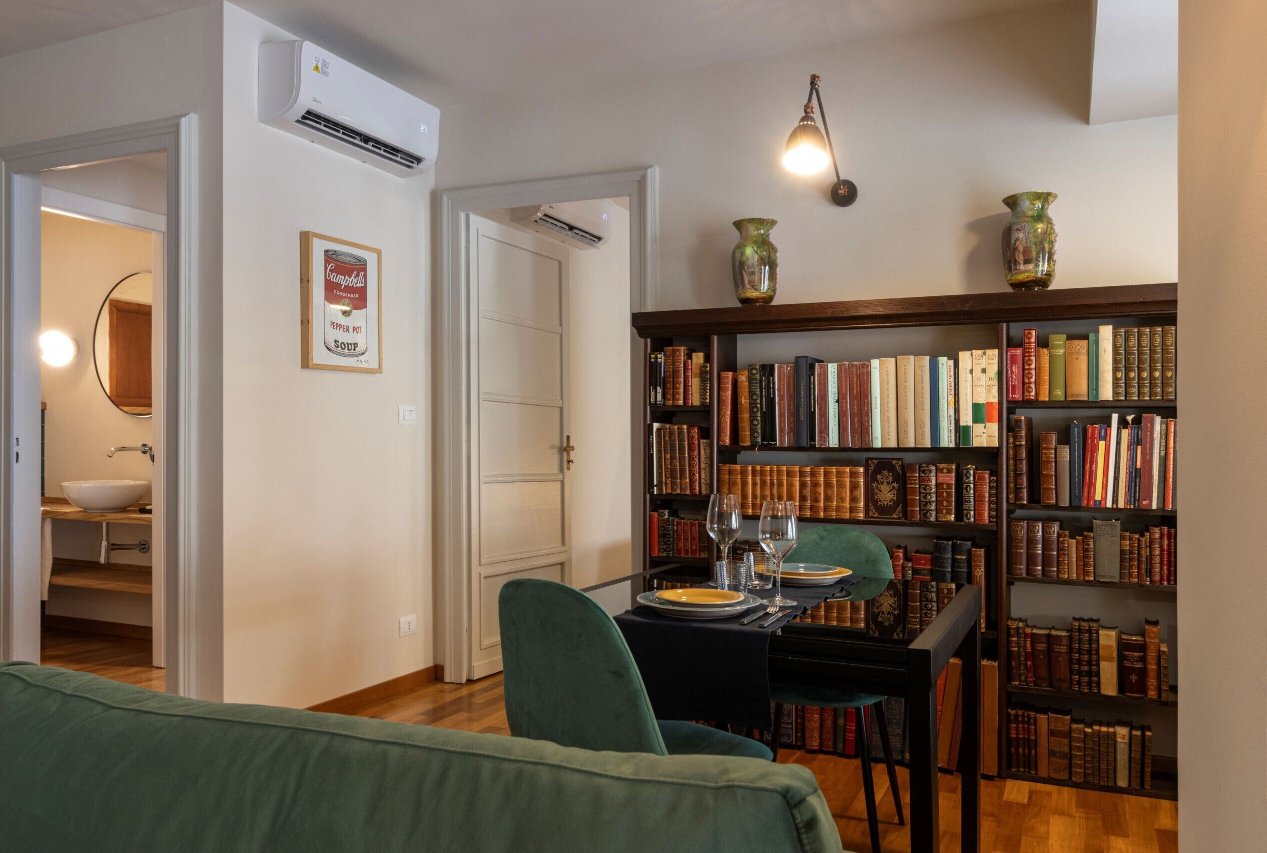A room with a bookshelf, a dining table with chairs, a green sofa, and a small kitchen area with a sink and a bowl.