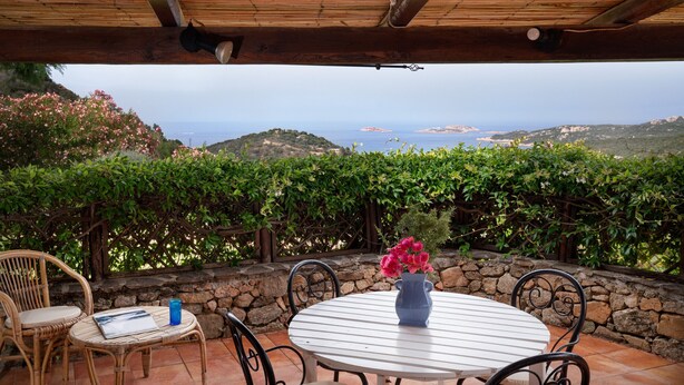 An outdoor patio with a table and chairs, a stone wall, and a view of the sea and hills. - Casa del Bonsai by Wonderful Italy (Arzachena)