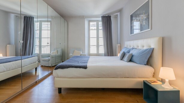 A modern bedroom with a large bed, a bedside table, a mirror, and a view of the outdoors through large windows. - Vittorio Place by Wonderful Italy (Turin)