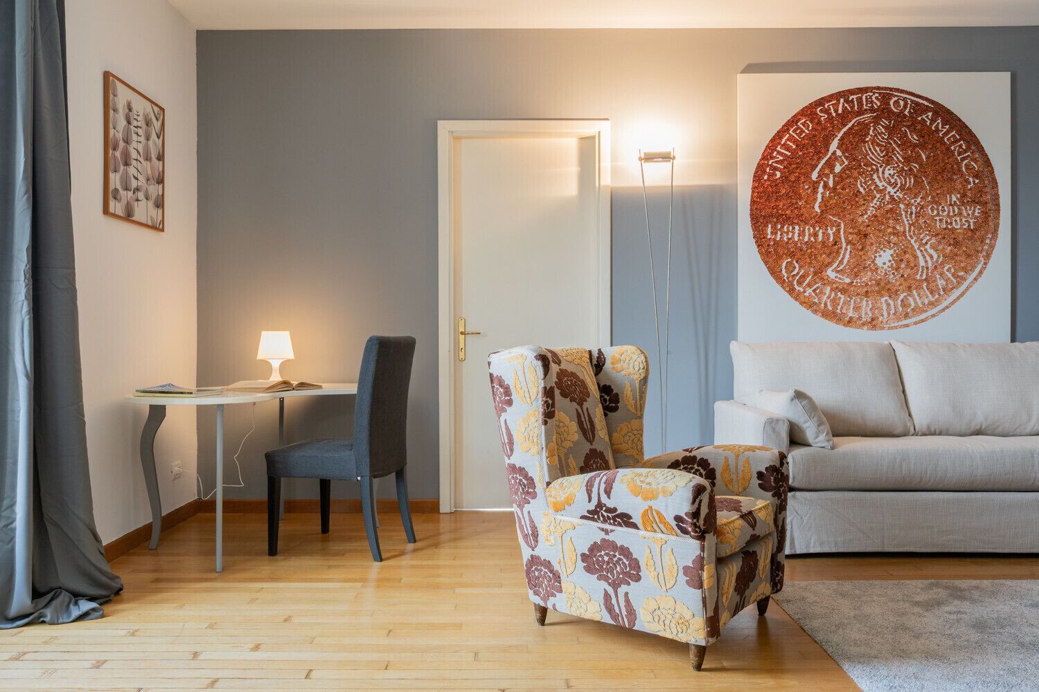 A living room with a sofa, a chair, a desk with a lamp, and a wall with a large coin design.