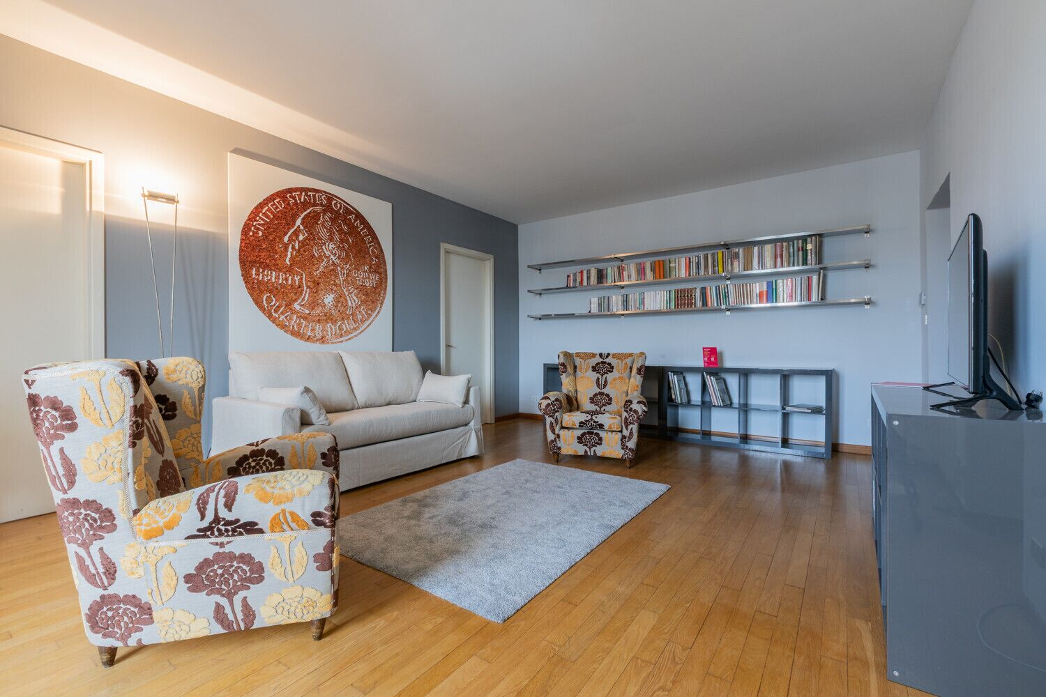 A modern living room with a sofa, two patterned armchairs, a coffee table, a bookshelf, and a television.