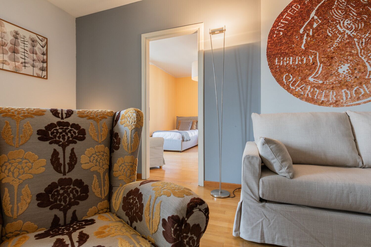 A living room with a sofa, a patterned armchair, a floor lamp, and a bed in the background.