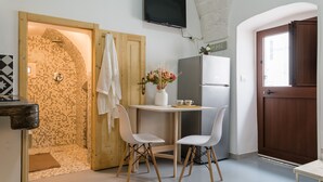 Une cuisine compacte avec un mur recouvert de carreaux, une porte en bois, un réfrigérateur blanc et une petite table accompagnée de deux chaises blanches.