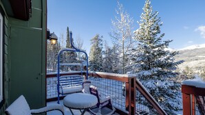 Un balcone imbiancato dalla neve con una sedia e un tavolo, affacciato su un paesaggio innevato con alberi e una montagna sullo sfondo.