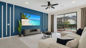 Ein modernes Wohnzimmer mit großem Fernseher, Deckenventilator und Blick auf einen Pool durch große Glasüren.