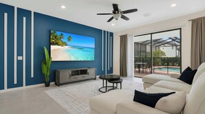 A modern living room with a large TV, a ceiling fan, and a view of a pool through large glass doors. - Cozy 5BR Solterra Home w/ Private Pool (Davenport)