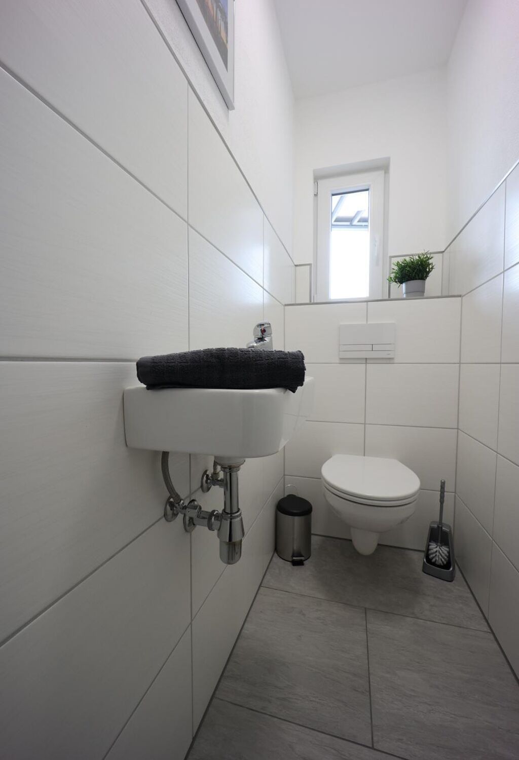 Ett litet, rent badrum med ett vitt handfat, en toalett och ett litet fönster ovanför toaletten.