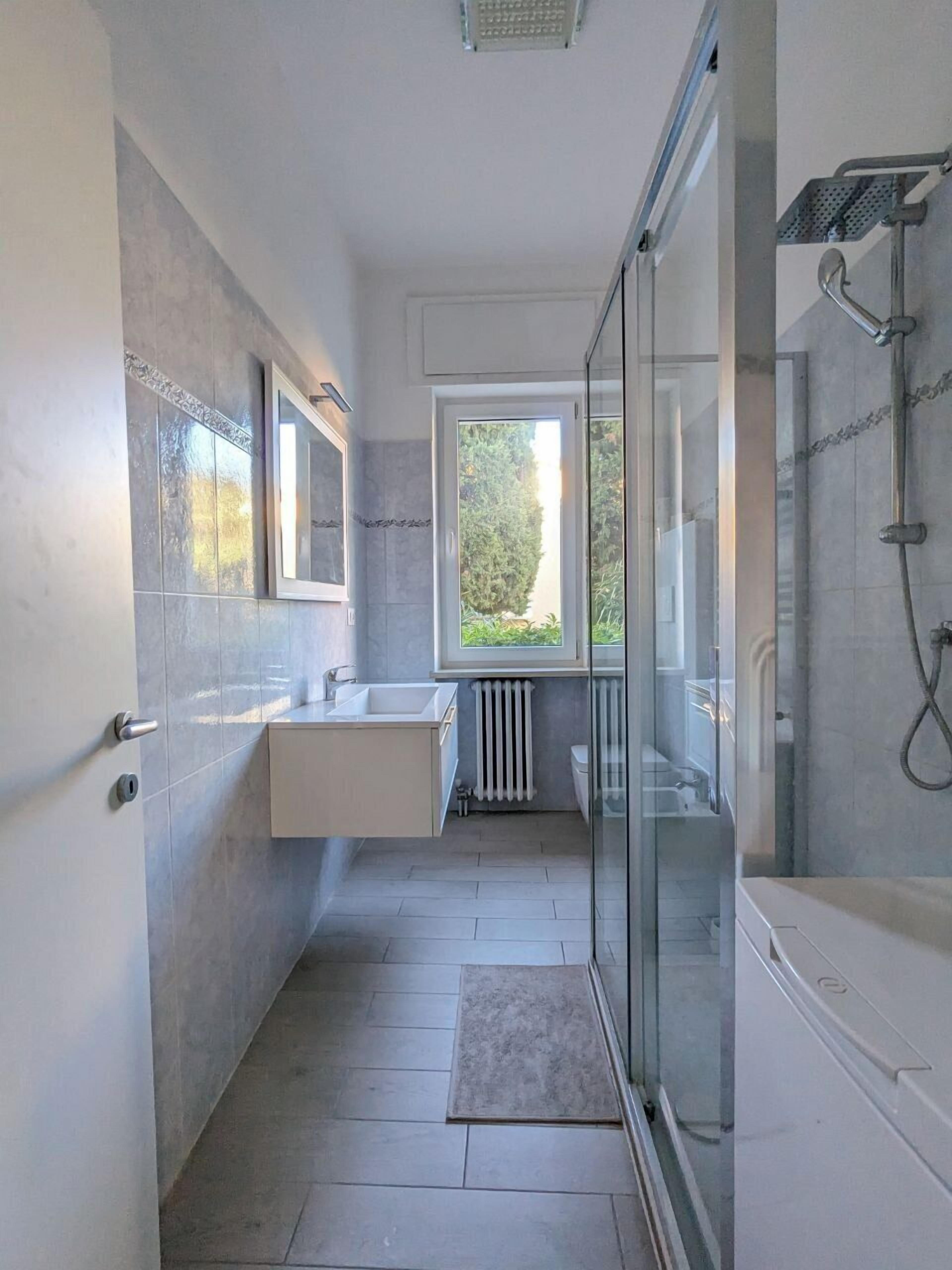 Ein modernes Badezimmer mit Duschkabine aus Glas, einem weißen Waschbecken und einem Fenster, das für natürliches Licht sorgt.