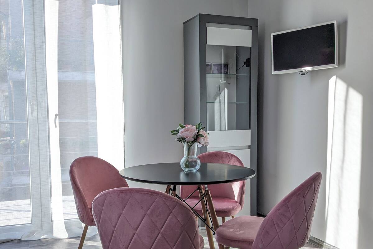 Un comedor moderno con una mesa redonda, cuatro sillas rosadas, un florero con flores, un espejo grande y un televisor de pantalla plana montado en la pared.