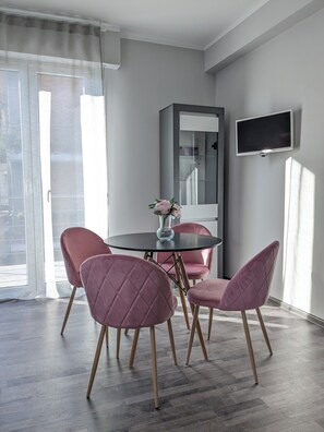 Un espace repas moderne avec une table ronde, quatre chaises roses, un vase avec des fleurs, un grand miroir et un téléviseur à écran plat fixé au mur.