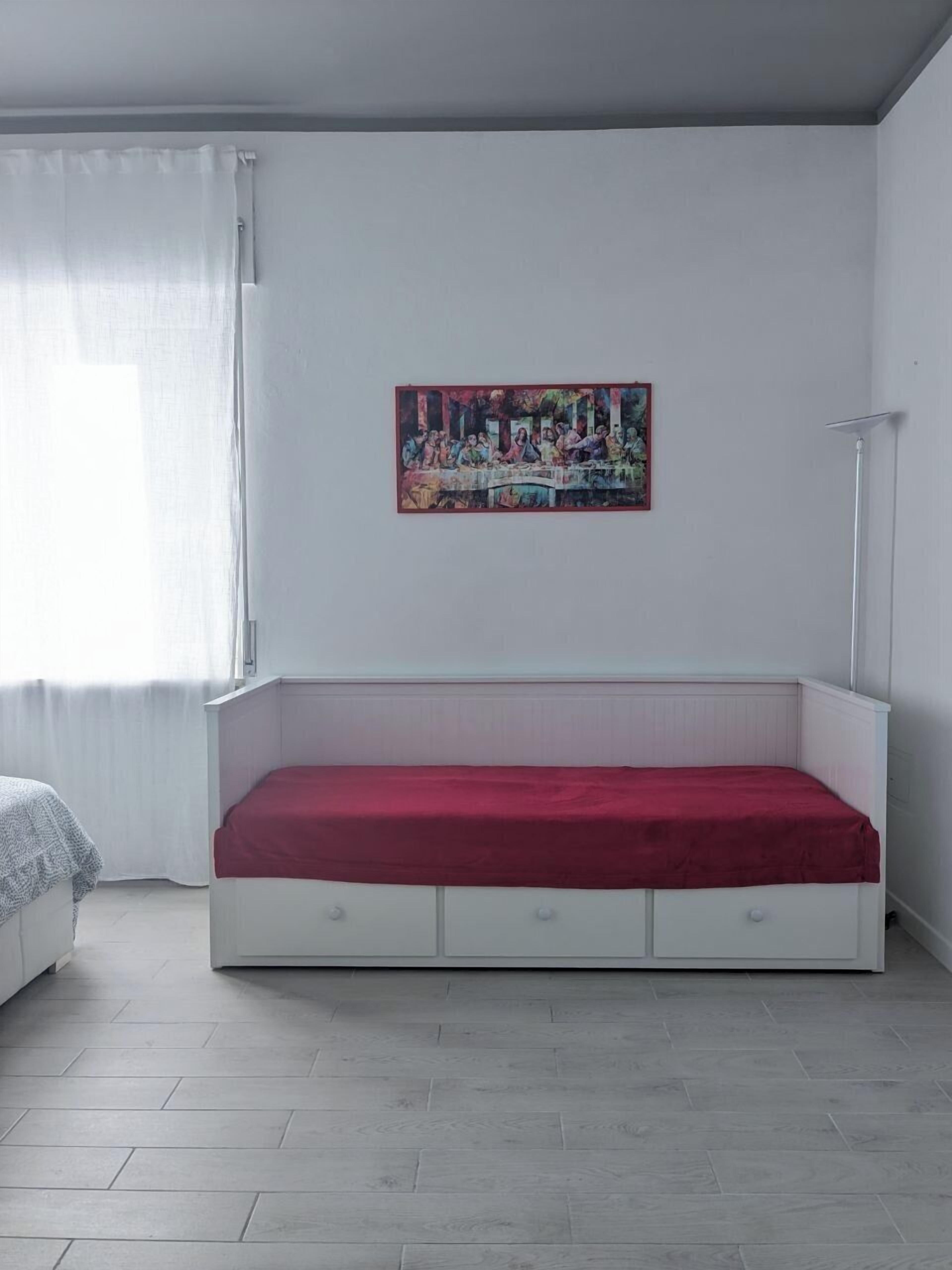 A minimalist bedroom with a white bed, a colorful artwork on the wall, and a white bedside table.