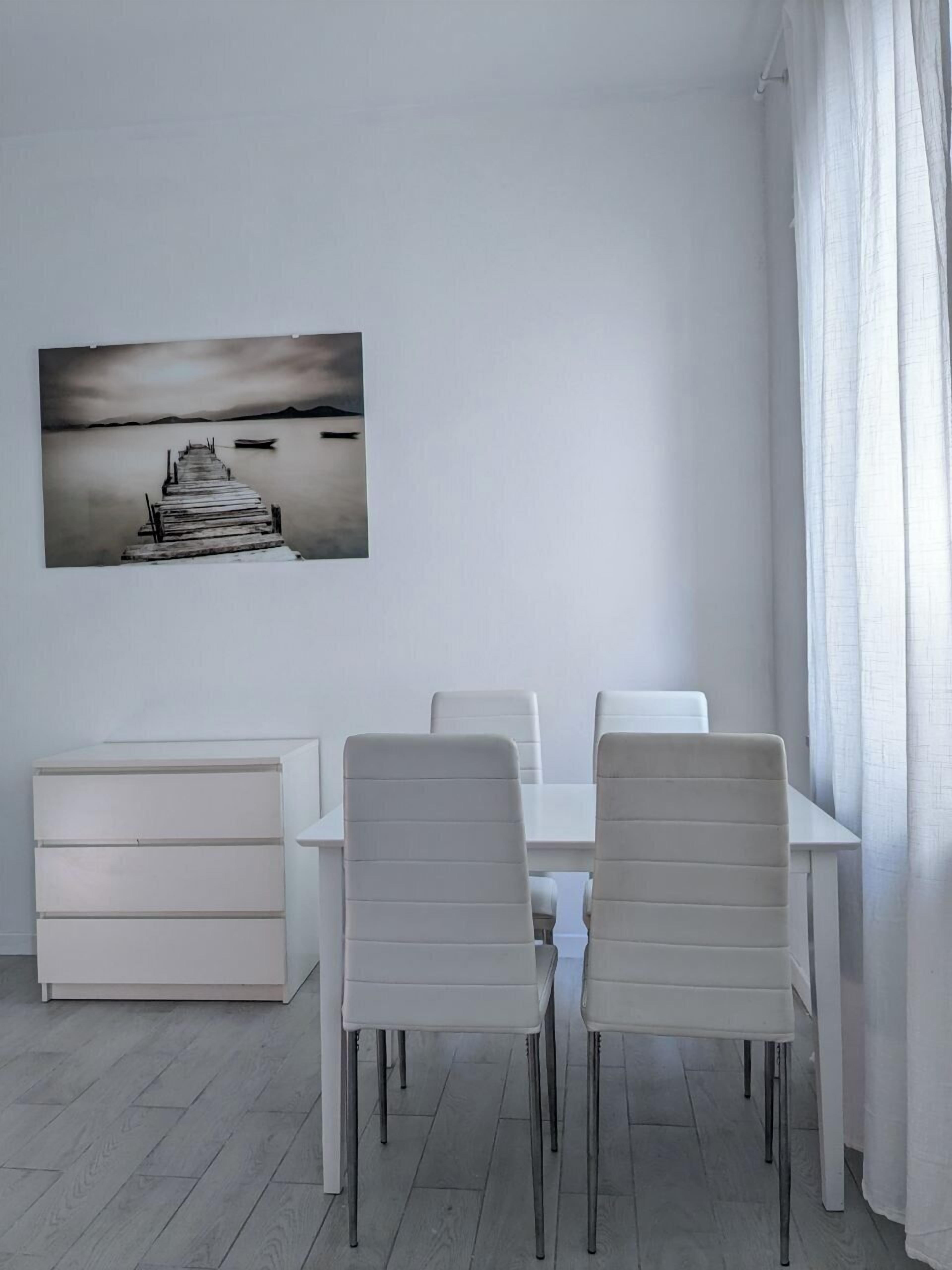 A modern dining area with a white table and chairs, a white cabinet, and a large black and white painting of a pier.