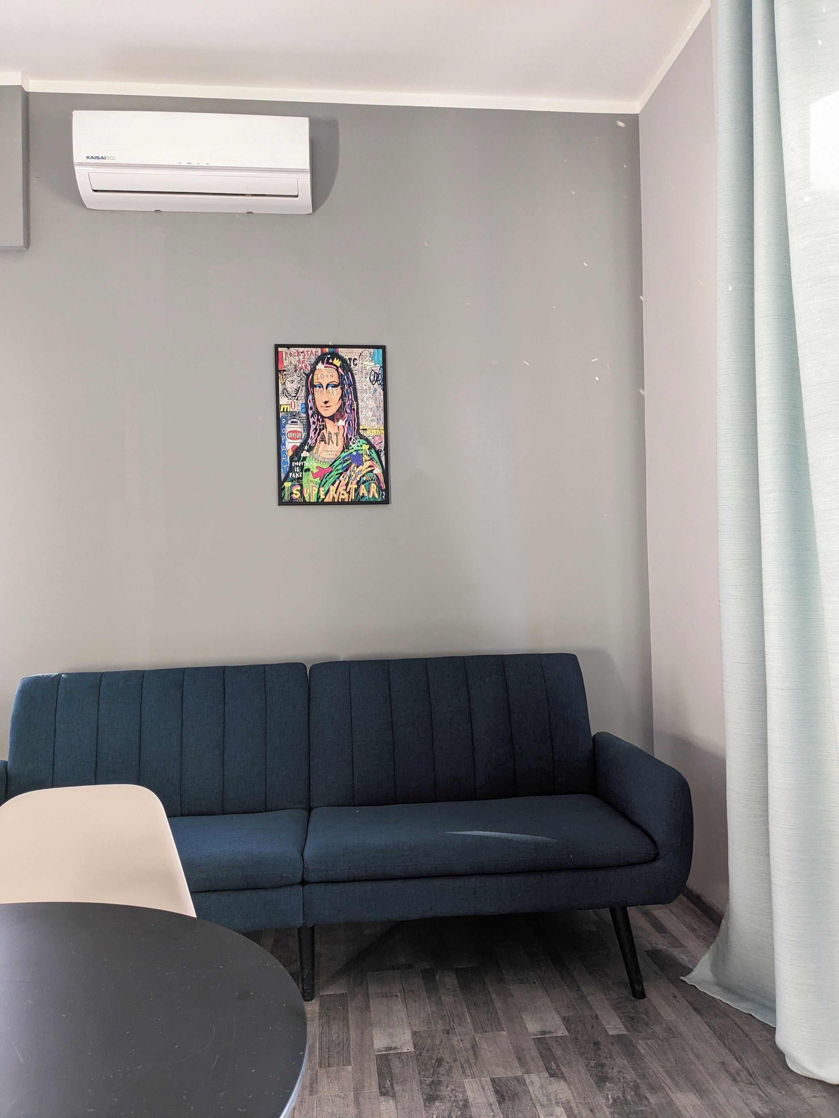A room with a dark blue sofa, a round table, an air conditioning unit, and a colorful artwork on the wall.