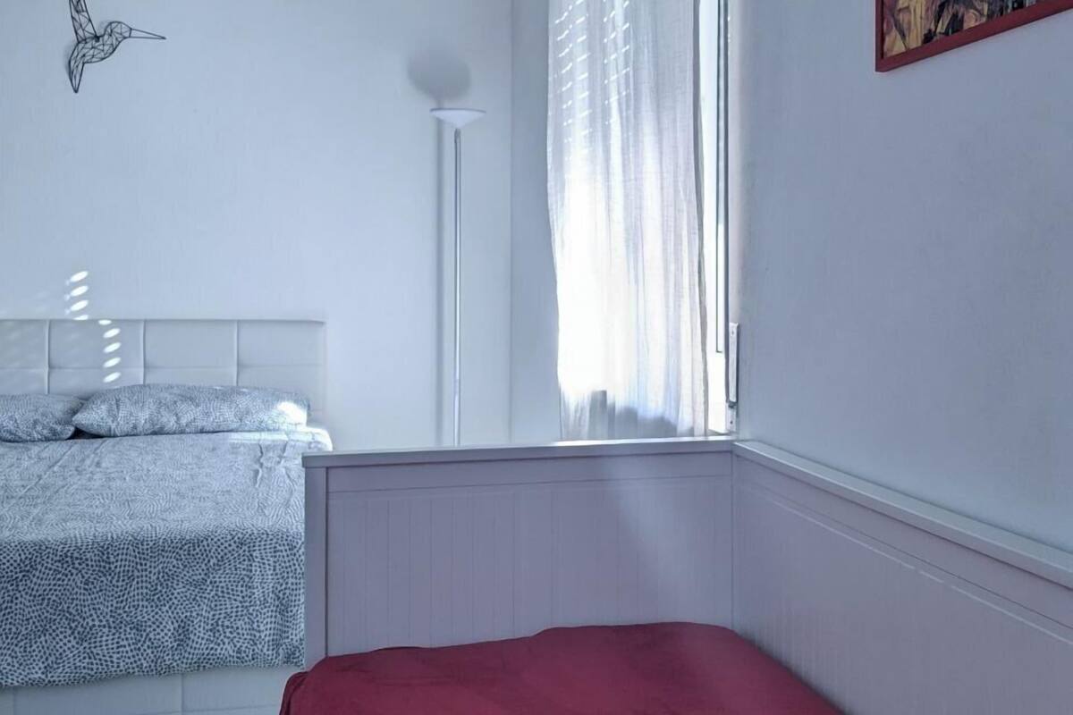 Un dormitorio con una cama, una colcha roja, una pared blanca, una ventana con cortinas blancas y un cuadro.