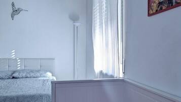 Un dormitorio con una cama, una colcha roja, una pared blanca, una ventana con cortinas blancas y un cuadro.