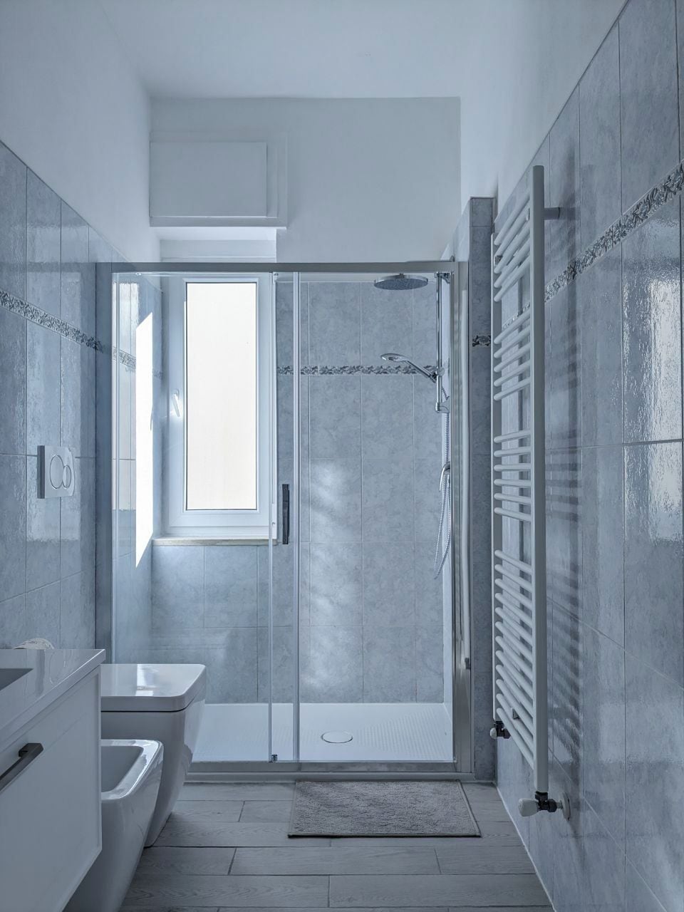 Un baño moderno con cabina de ducha de vidrio, un lavabo blanco y un ventanal que proporciona luz natural.