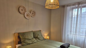 Un dormitorio con una cama, dos almohadas, una lámpara de noche, una lámpara colgante y una ventana con cortinas.