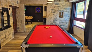 Una mesa de billar con bolas, un dardo y una silla de madera en una habitación con paredes de piedra y una ventana.