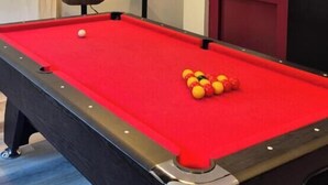 Una mesa de billar con superficie de fieltro rojo, tres lámparas colgantes negras encima y una puerta con panel de vidrio esmerilado a la derecha.