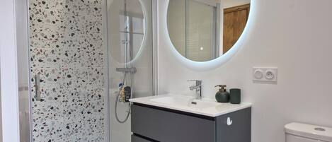 A modern bathroom with a glass shower enclosure, a round mirror, a white sink, and a dark vanity.