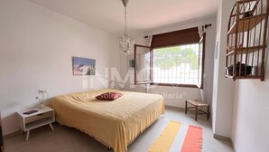 Ein Schlafzimmer mit Bett, Nachttisch, Lampe, Fenster und einem kleinen Hocker.
