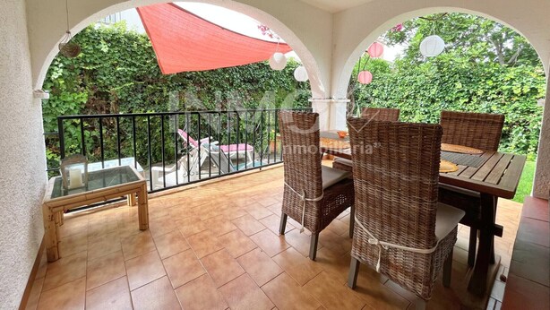 Ein überdachter Patio-Bereich mit Tisch und Stühlen, umgeben von einem schwarzen Metallgeländer und Blick auf Grünflächen.