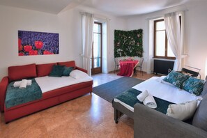 Une pièce avec un canapé rouge, un lit avec une literie verte, une petite table recouverte d’une nappe rouge et un mur orné d’un motif floral.