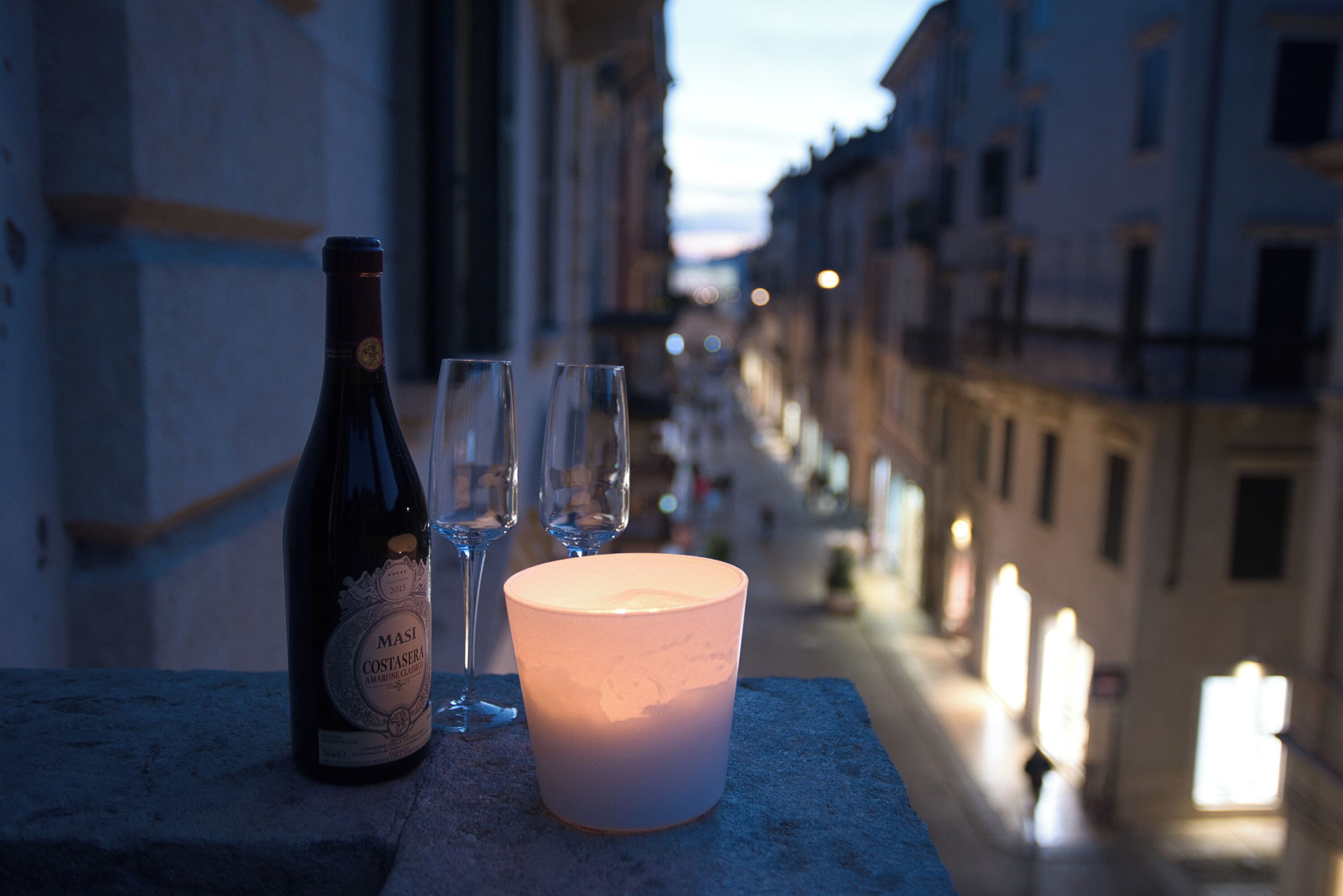 A bottle of Masi Costasera wine, two glasses, and a candle on a stone ledge overlooking a street at dusk.