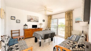 A room with a dining table, chairs, a sofa, a TV, and a balcony with curtains. - Apartament with barbecue in La Llosa (Cambrils)