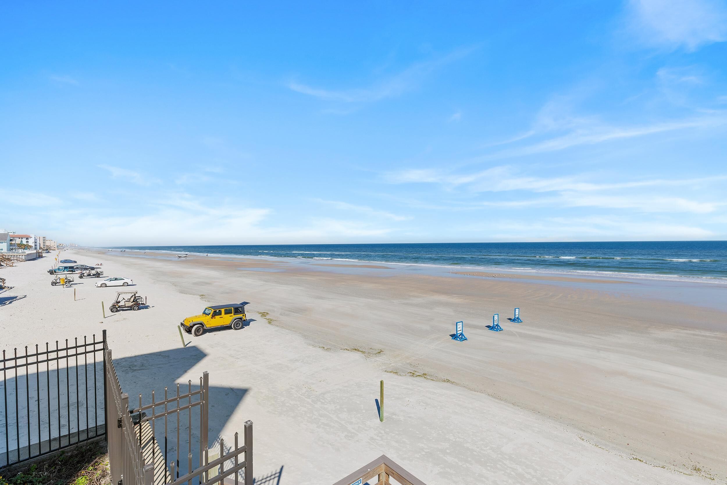 Ein Strand mit einem gelben Fahrzeug in Strandnähe, einem Zaun und einigen Liegestühlen.
