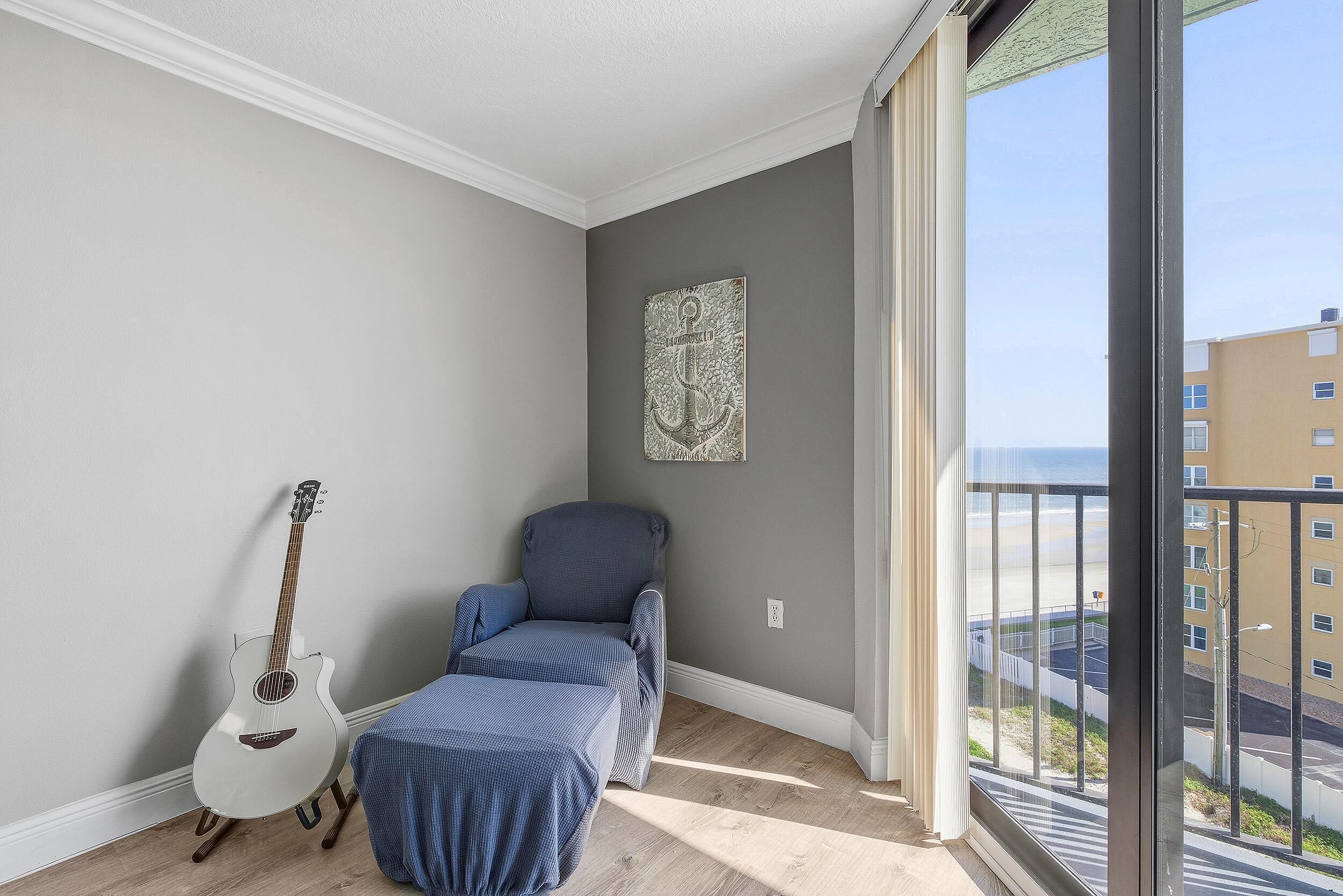 Ein Zimmer mit grauer Wand, einer weißen Gitarre, einem blauen Sessel und Blick auf den Strand durch ein großes Fenster.