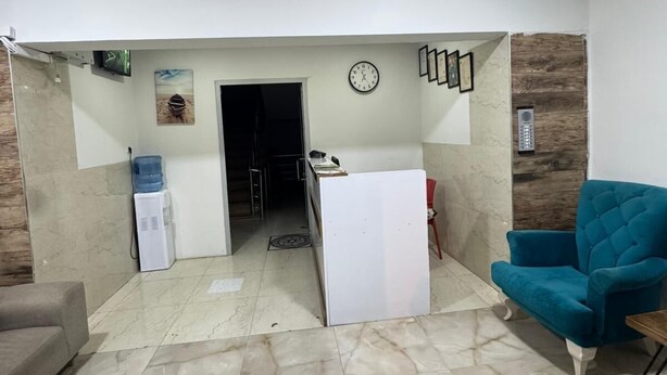 A spacious living area with a marble floor, a blue armchair, a sofa, a water dispenser, and a clock on the wall. - Acar apart otel (Antalya)