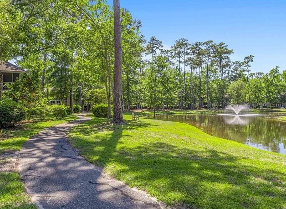 A pathway leading to a body of water with a fountain, surrounded by trees and grass.
