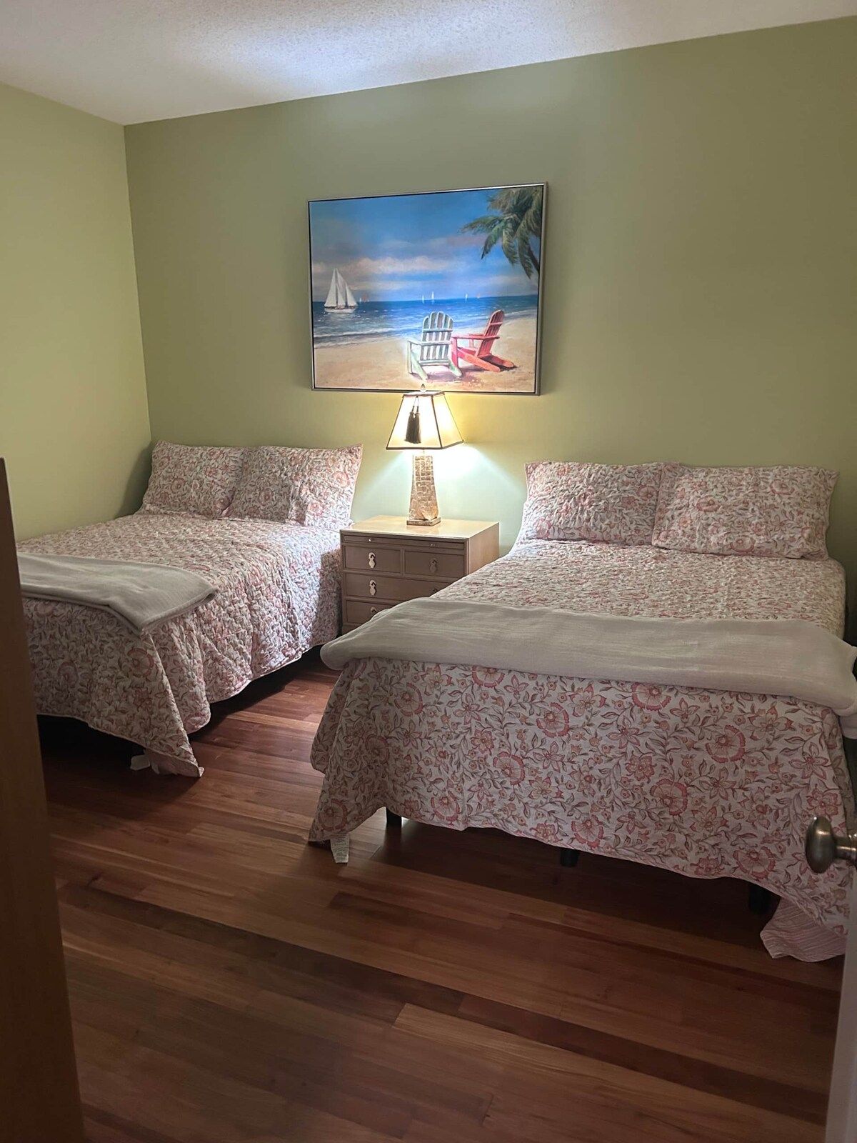 A bedroom with two beds, a nightstand with a lamp, and a painting of a beach scene on the wall.