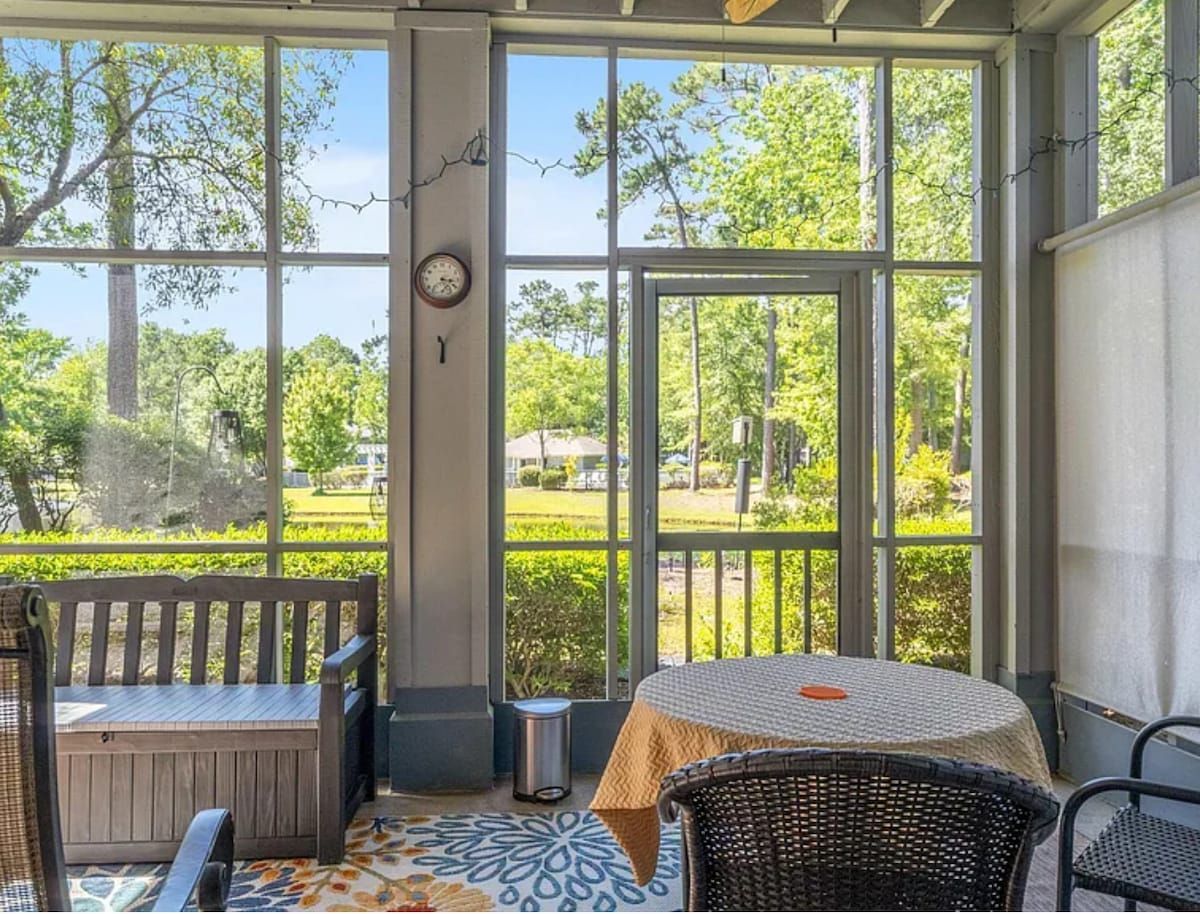 A covered outdoor seating area with a table and chairs, a bench, and a view of a green landscape through large windows.