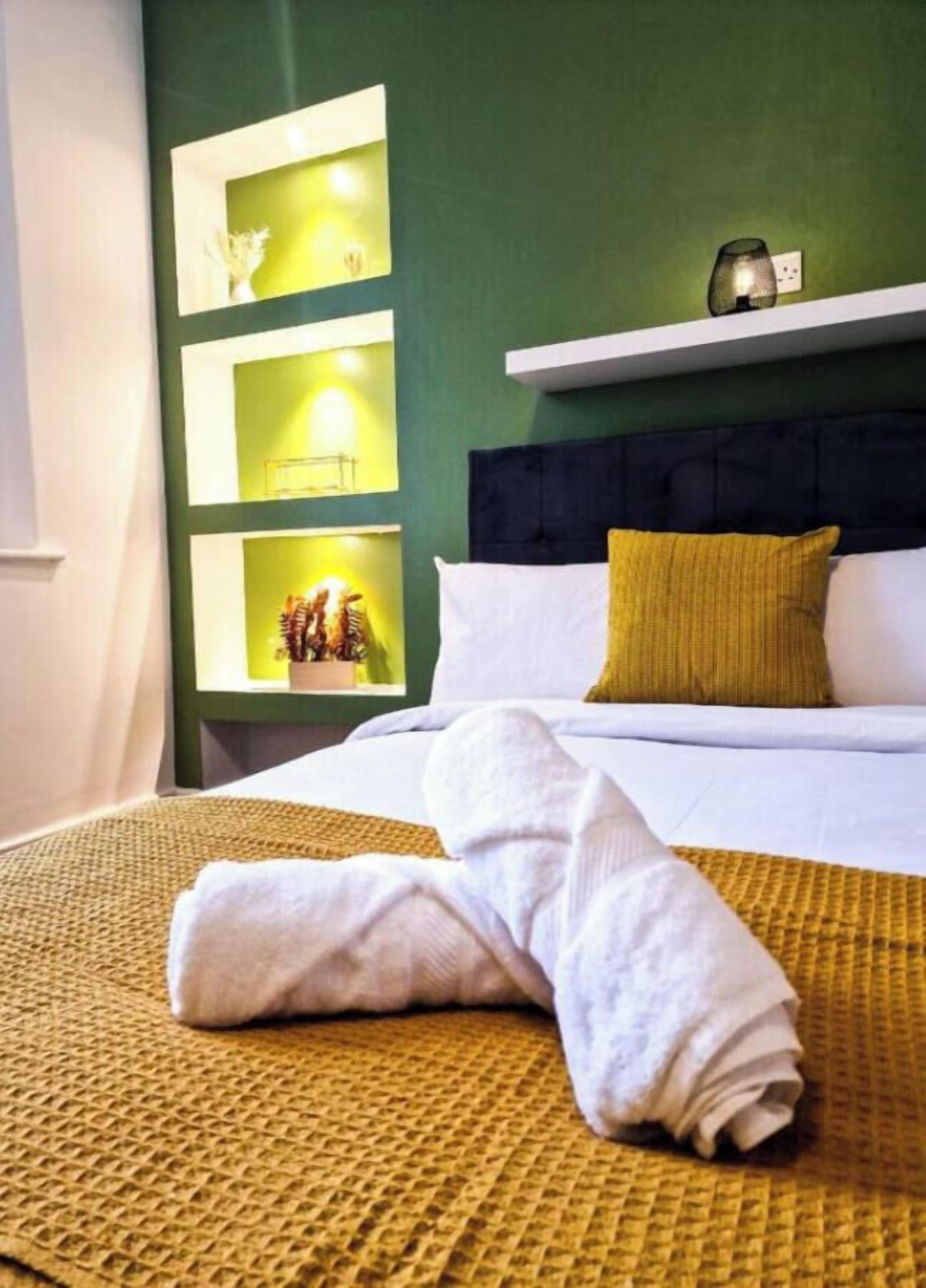 Una cama bien hecha con sábanas blancas y un plaid amarillo, cabecera oscura y paredes verdes con estantes que exhiben objetos decorativos.