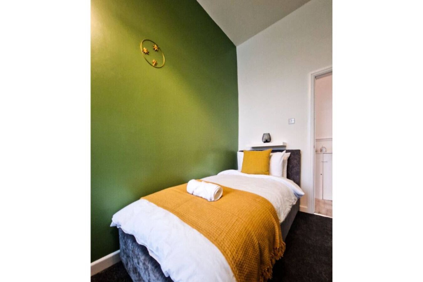 A bedroom with a green wall, a bed with white and mustard bedding, a nightstand with a lamp, and a door to the right.