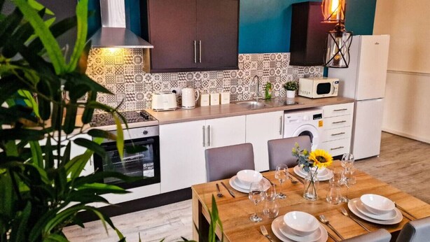 A modern kitchen with white cabinets, a patterned backsplash, and a dining area set for six with a table, chairs, and a vase of sunflowers.