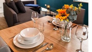 A dining area with a wooden table set for four, a TV mounted on the wall, and a sofa in the background.