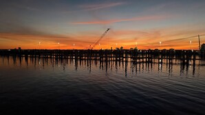 Eine Uferlandschaft im Abendlicht mit einem Steg, der sich in ruhiges Wasser erstreckt, welches den Himmel spiegelt.