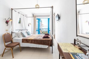 Una habitación con cama, una silla, una mesa, televisor y balcón con barandilla azul.