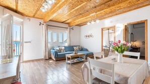 A spacious living area with a wooden ceiling, a blue sofa, a dining table with chairs, and a view through a large window.