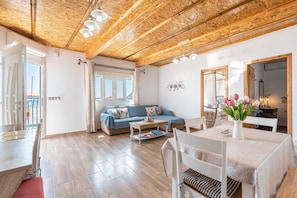 A spacious living area with a wooden ceiling, a blue sofa, a dining table with chairs, and a view through a large window.