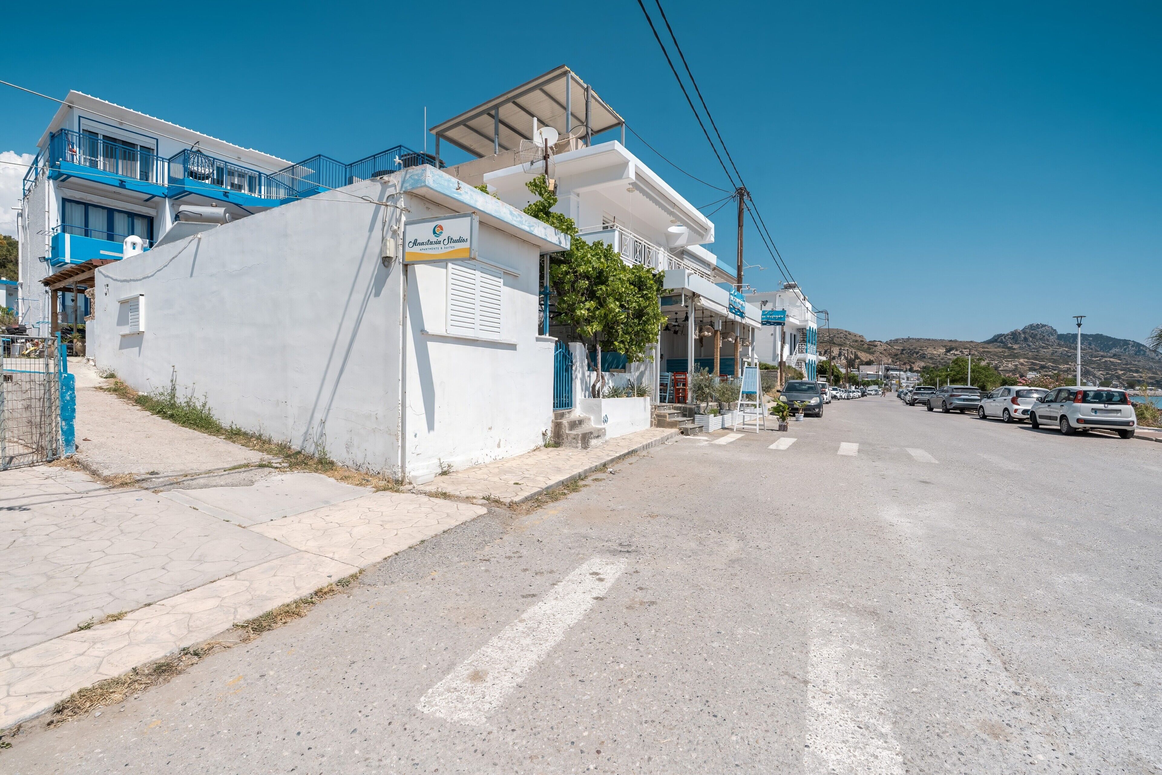 A coastal street with white buildings, a blue balcony, and a clear sky.