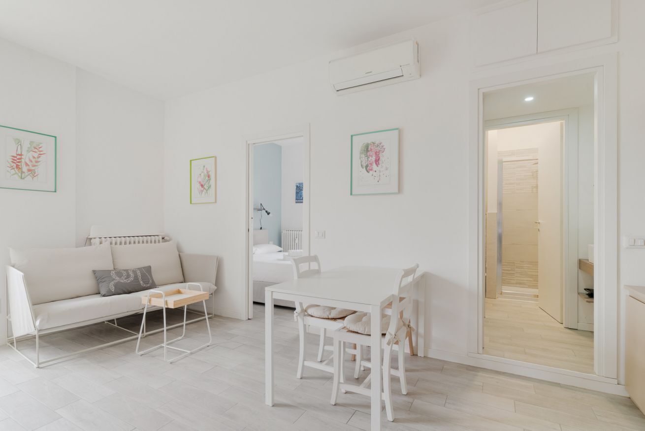 A modern, minimalist living room with a sofa, a dining table, and a kitchen visible through an open door.