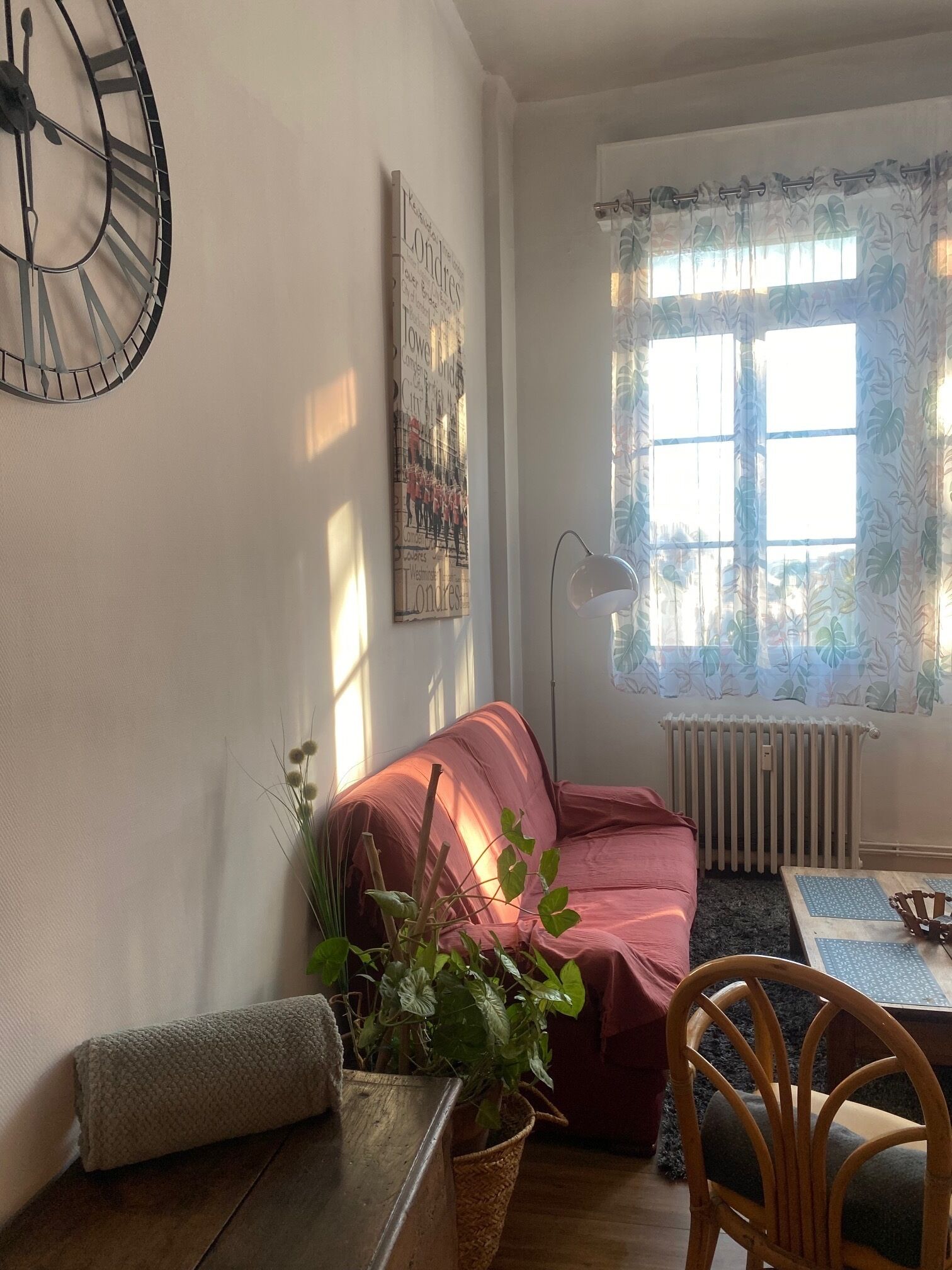 A cozy living room with a red sofa, a wooden coffee table, a wicker side table, a potted plant, a wooden chair, a wall clock, a poster, a window with sheer curtains, and a radiator.
