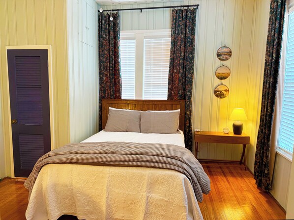 Um quarto com cama, mesa de cabeceira com abajur, porta e cortinas.