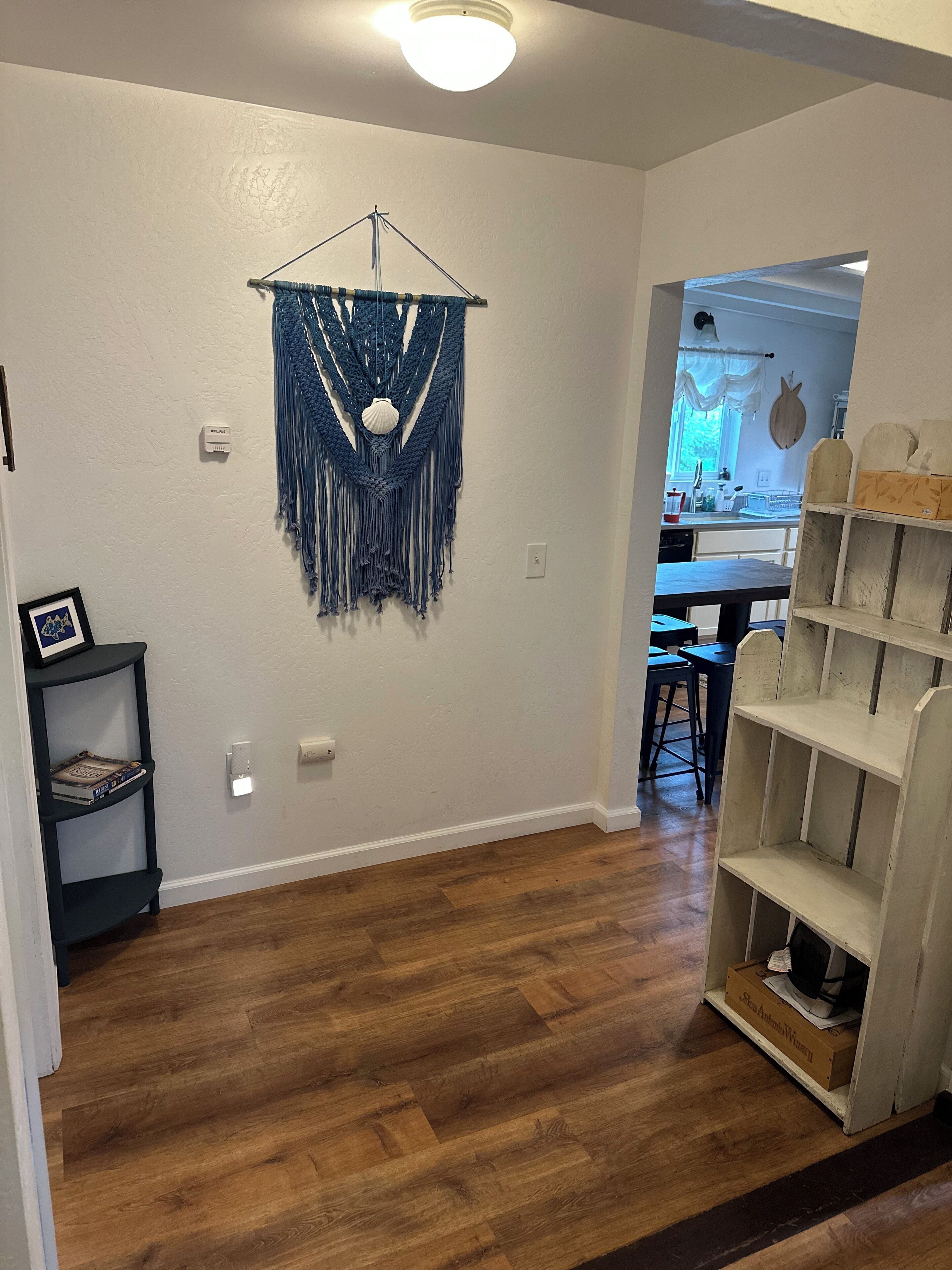 A room with a wooden herringbone floor, a macrame wall hanging, a small shelf with books, and a wooden shelf unit.