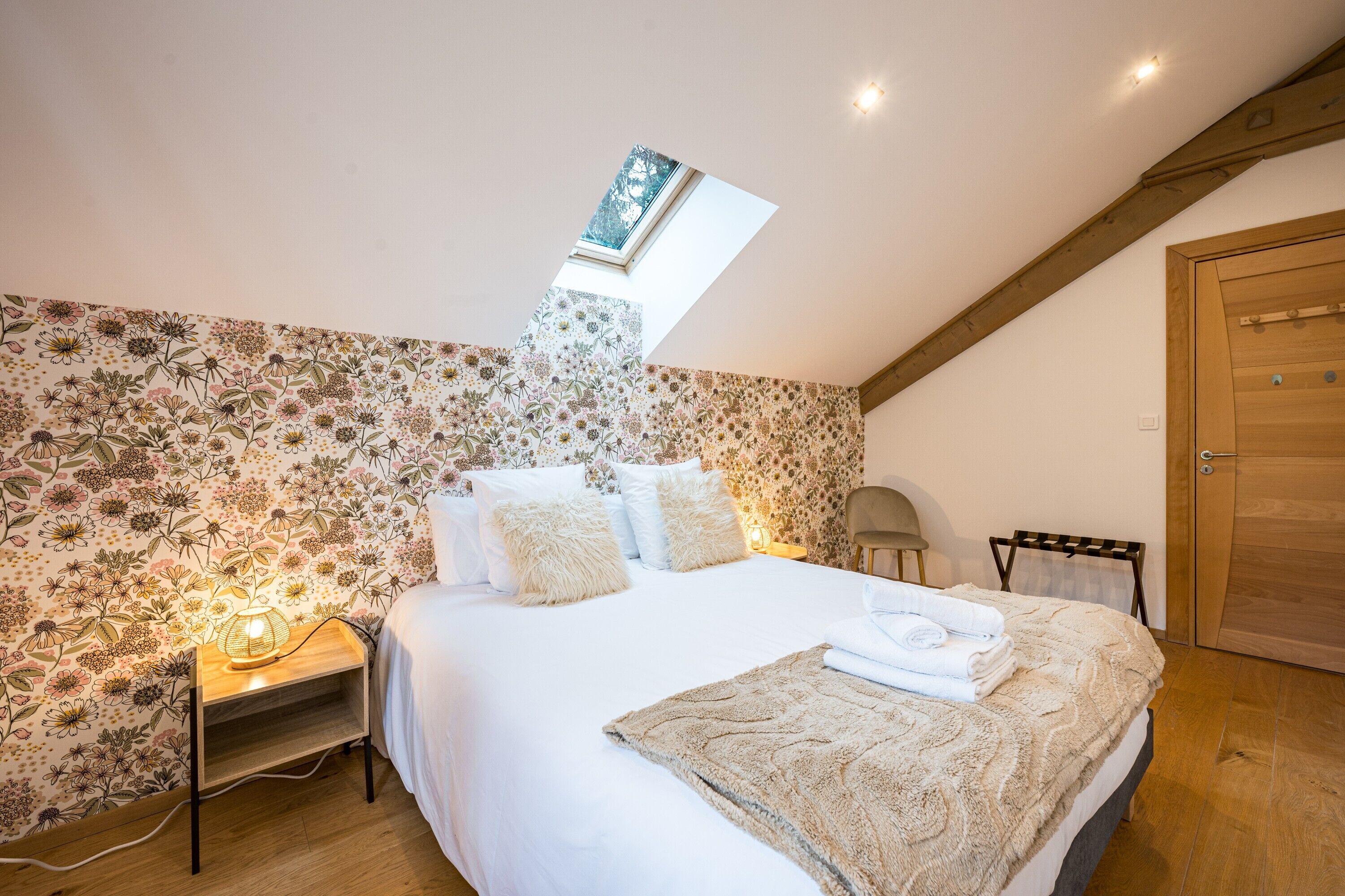 A bedroom with a skylight, floral wallpaper, a bed with white bedding, a bedside lamp, and a wooden wardrobe.