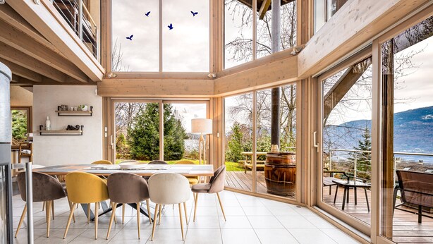 Un vaste espace dédié à la salle à manger, avec une grande table et des chaises, une vue sur l’extérieur grâce à de grandes fenêtres, et un intérieur en bois.